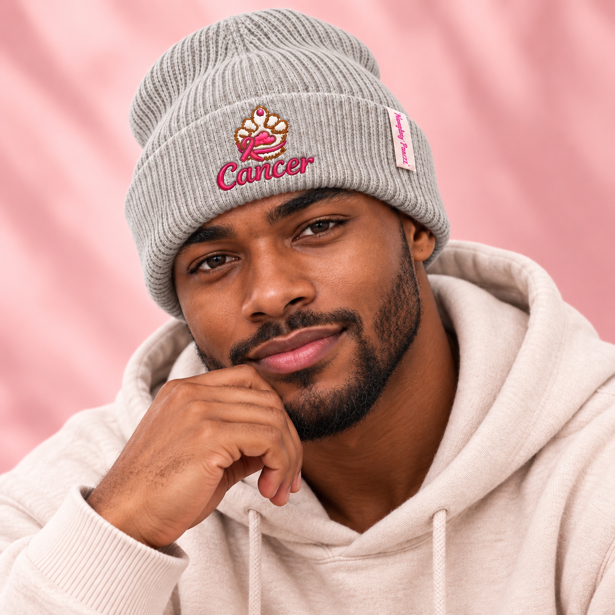 A man wearing a light gray ribbed knit beanie with a pink and gold Cancer zodiac sign embroidery, paired with a cream-colored hoodie against a soft pink background.