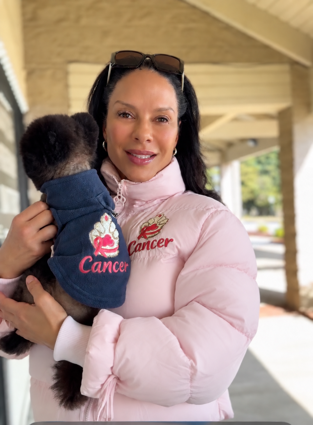 Women is a pink puffer jacket with an embroidered  paw logo with the word cancer underneath the paw holding a dog with a blue fleece with an embroidered paw logo with a pink ribbon around the paw with the word cancer underneath the paw.