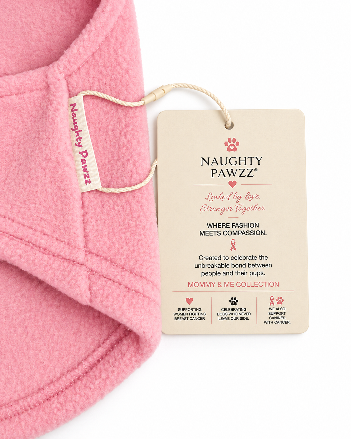 Close-up of a soft pink fleece pet sweater with an embroidered paw print, pink ribbon, and the word 'Cancer' in pink. A beige hangtag for Naughty Pawzz is attached near the collar.