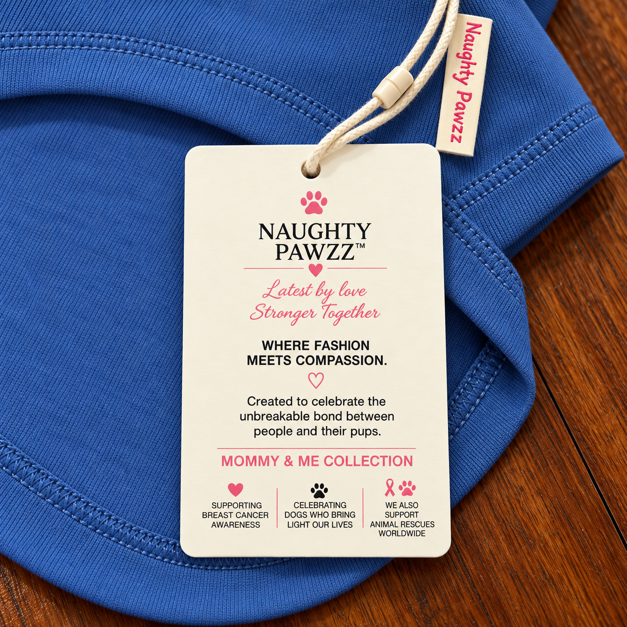 A close-up of a folded blue garment with a hanging paper brand tag reading Naughty Pawzz and a small side tag also showing the brand, placed on a wooden surface.