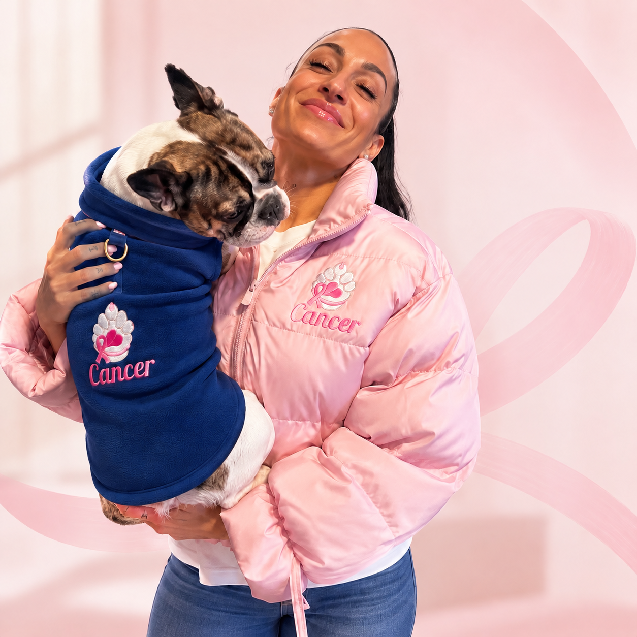 Women is a pink puffer jacket with an embroidered  paw logo with the word cancer underneath the paw holding a dog with a blue fleece with an embroidered paw logo with a pink ribbon around the paw with the word cancer underneath the paw.
