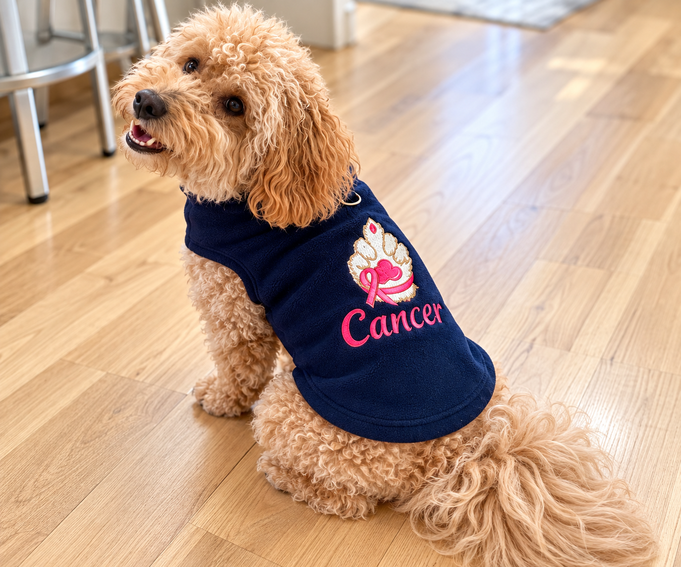 A small tan dog wearing a navy blue pet shirt with a pink ribbon and the word 'Cancer' on the front, sitting on a light wood floor in an indoor setting.