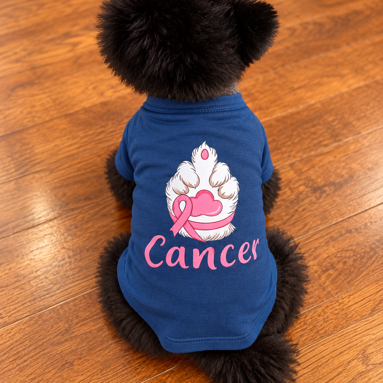 A small black dog viewed from behind wearing a blue pet shirt with a paw print, pink ribbon, and the word 'Cancer' printed on the back, sitting on a wooden floor.