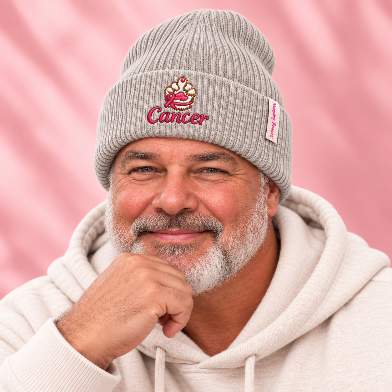 A smiling man with a gray beard wearing a light gray ribbed beanie with a pink and gold crab logo and the word 'Cancer' embroidered on the front, paired with a cream hoodie against a soft pink background.