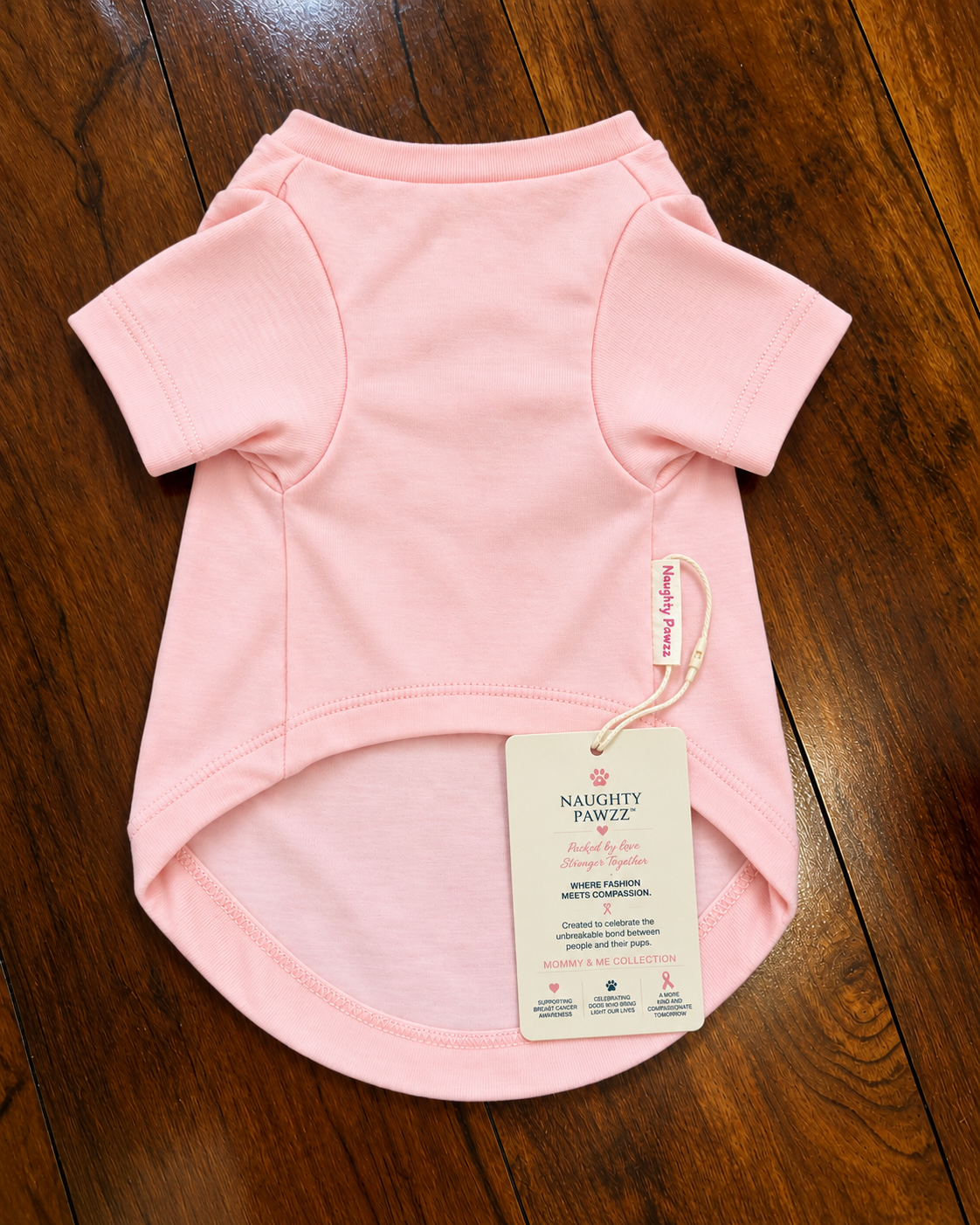 A light pink short-sleeve dog shirt laid flat on a dark wood floor, with a hangtag attached. The tag reads Naughty Pawz.