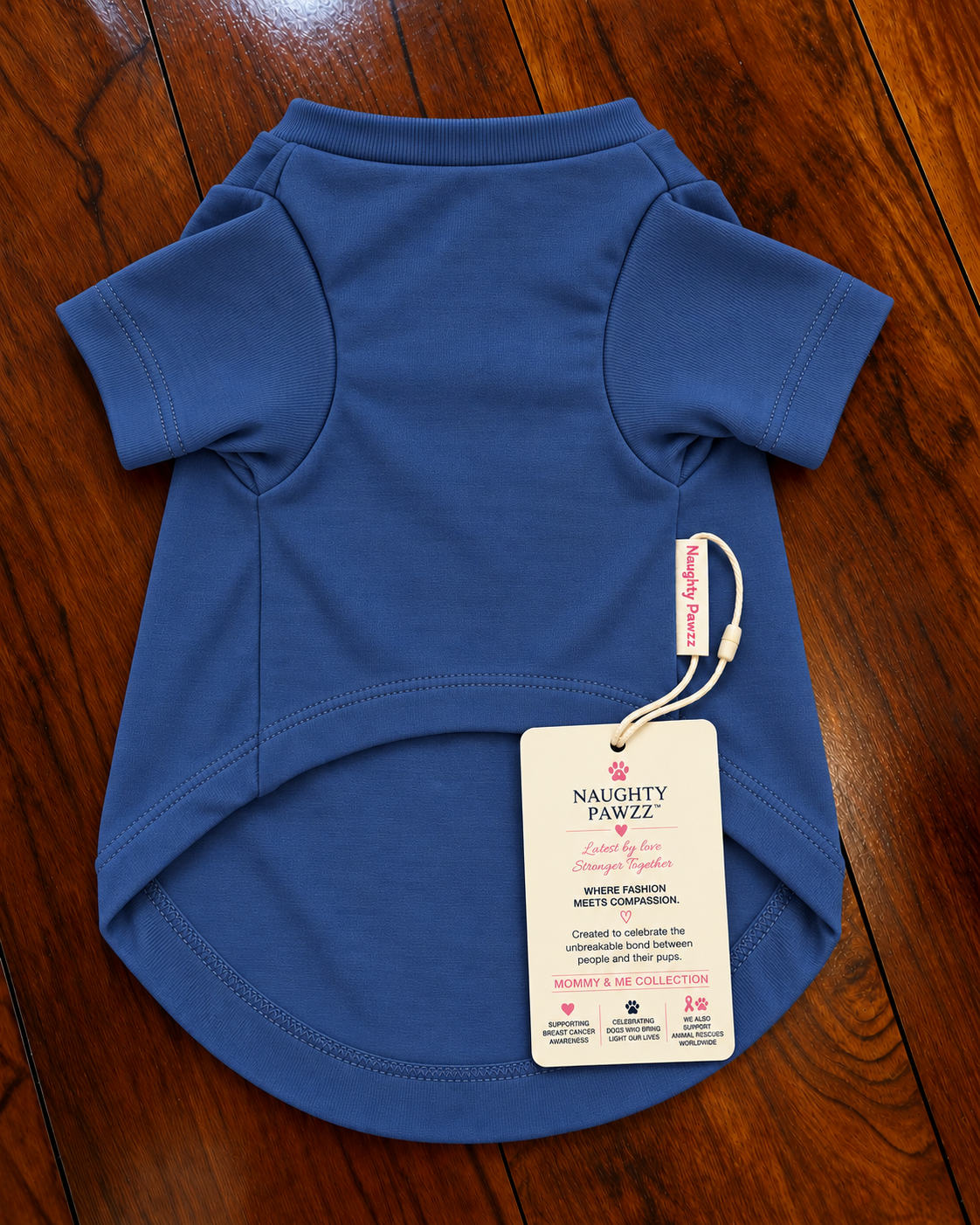 A royal blue short-sleeve dog t-shirt laid flat on a dark wood floor, featuring a white hangtag with 'Naughty Pawz' branding.