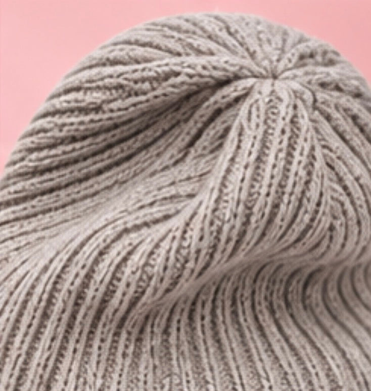 A close-up of a ribbed knit beanie hat in a light taupe-gray color, set against a solid pink background.